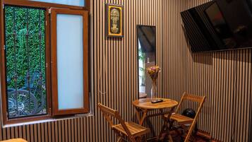 Una habitación con piso de madera, ventana con vista a follaje, una pequeña mesa con sillas y un televisor montado en la pared.