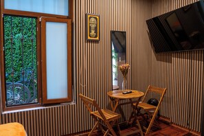 A room with a wooden floor, a window with a view of greenery, a small table with chairs, and a television mounted on the wall. - Posada Miraflores (Lima)