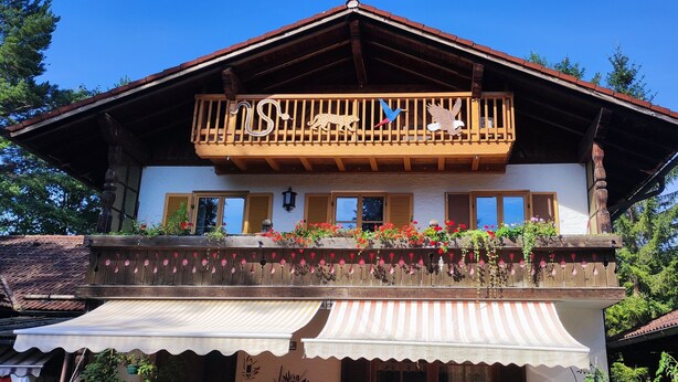 A traditional chalet with a balcony featuring a wooden railing, a decorative animal and bird motif, and a flower box with red flowers. - Relaxation on 2 levels; hiking,cycling from the house, skiing in the surrounding (Großweil)