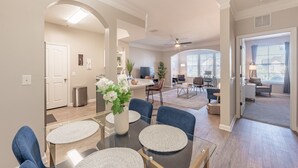 A spacious living area with a glass dining table, blue chairs, and a vase of flowers. There is a kitchen area in the background with a dining table and chairs.