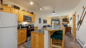 A kitchen with wooden cabinets, a refrigerator, a microwave, and a stove. There is a bar area with stools and a television.