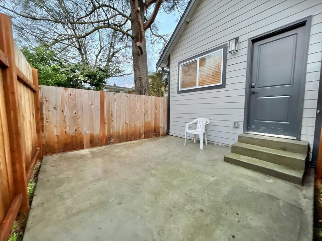 Un jardin avec une clôture en bois, une terrasse en béton, une chaise en plastique blanche et une maison grise avec une porte et une fenêtre.