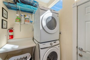 A laundry room with stacked washers and dryers, a fire extinguisher, and cleaning supplies on a shelf. - Gulf View * Sunsets * Boat parking * King Bed * Walk to Downtown (Cedar Key)