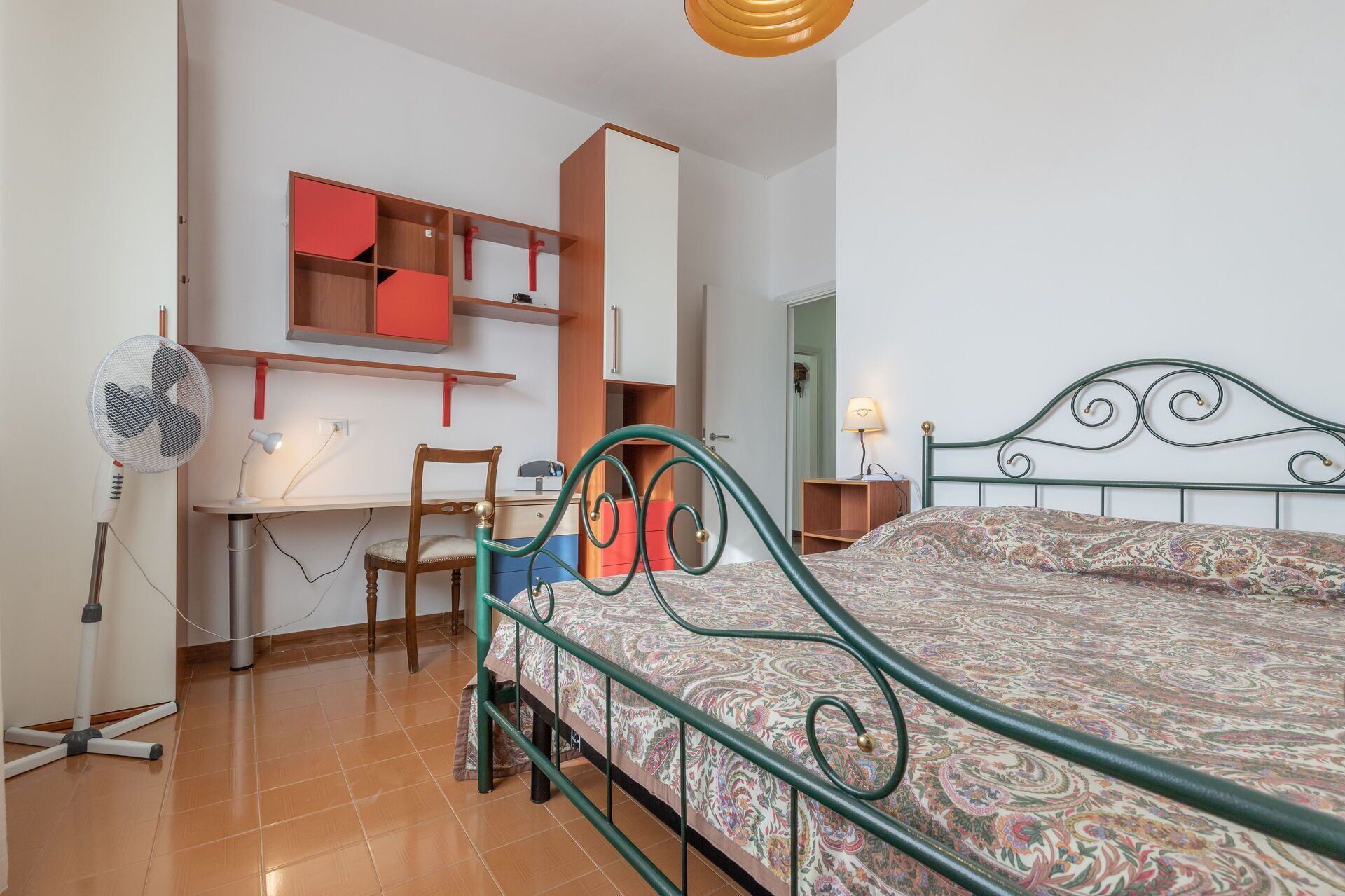 Een slaapkamer met een bed, bureau, stoel en een ventilator.