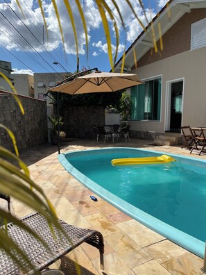 A swimming pool with a yellow inflatable float, surrounded by stone tiles and a covered patio area with chairs and an umbrella.