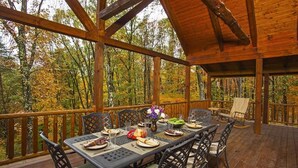 A covered outdoor dining area with a wooden deck, surrounded by trees with autumn foliage.