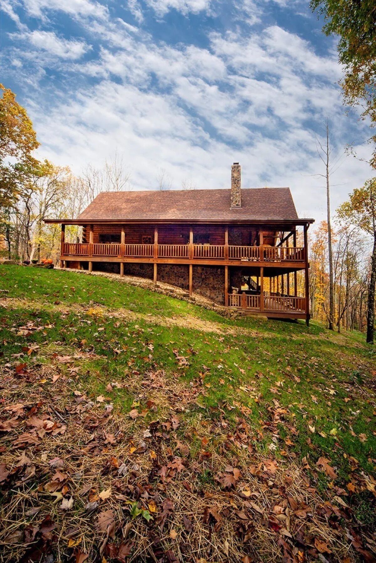 A wooden cabin with a stone chimney, a balcony, and a stone foundation, surrounded by a grassy area and trees.