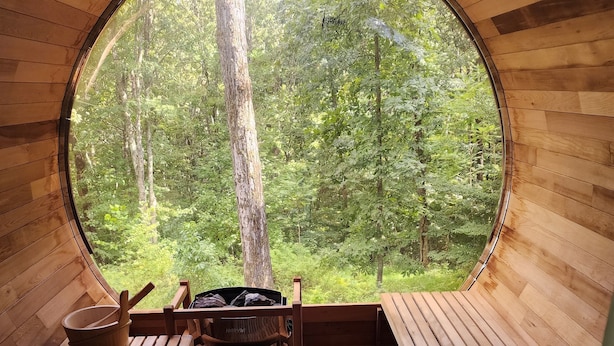 A circular wooden cabin with a large window, wooden benches, and a table with a fire pit.