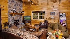 A cozy log cabin interior with a stone fireplace, wooden walls, a television, a sofa, a coffee table, and a window with a view of greenery.