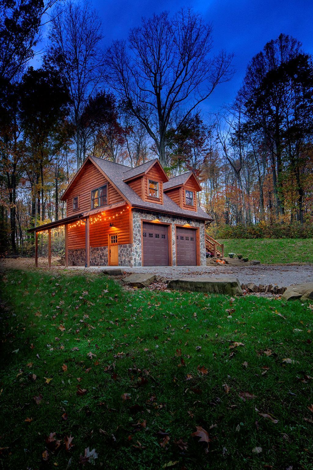 Une cabane en rondins avec des fondations en pierre, deux portes de garage et une véranda décorée de guirlandes lumineuses.