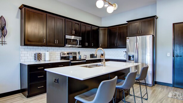 Una cocina moderna con armarios de madera oscura, electrodomésticos de acero inoxidable, una isla central con taburetes de bar y una lámpara decorativa en el techo.