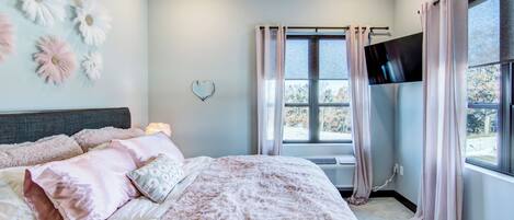 Une chambre à coucher avec un grand lit, un téléviseur à écran plat fixé au mur et une fenêtre avec des rideaux.