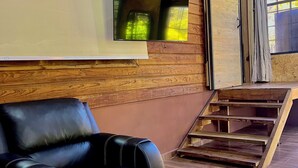 Quarto com sofá de couro preto, televisão de tela plana montada, paredes com painéis de madeira e escada de madeira.