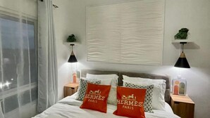 Una cama bien hecha con ropa blanca y dos almohadas rojas con el texto 'Hermès Paris'. Hay dos lámparas de noche, una ventana con cortinas traslúcidas y un estante en la pared con una planta.