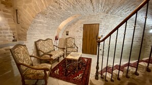 Una stanza con pareti in pietra, una scala in legno, due poltrone imbottite, un tavolino e un tappeto decorato.