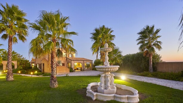 A landscaped garden with a fountain, palm trees, and a building with arched windows.