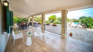 A covered outdoor patio with stone pillars, white chairs, and a table with a vase of flowers.