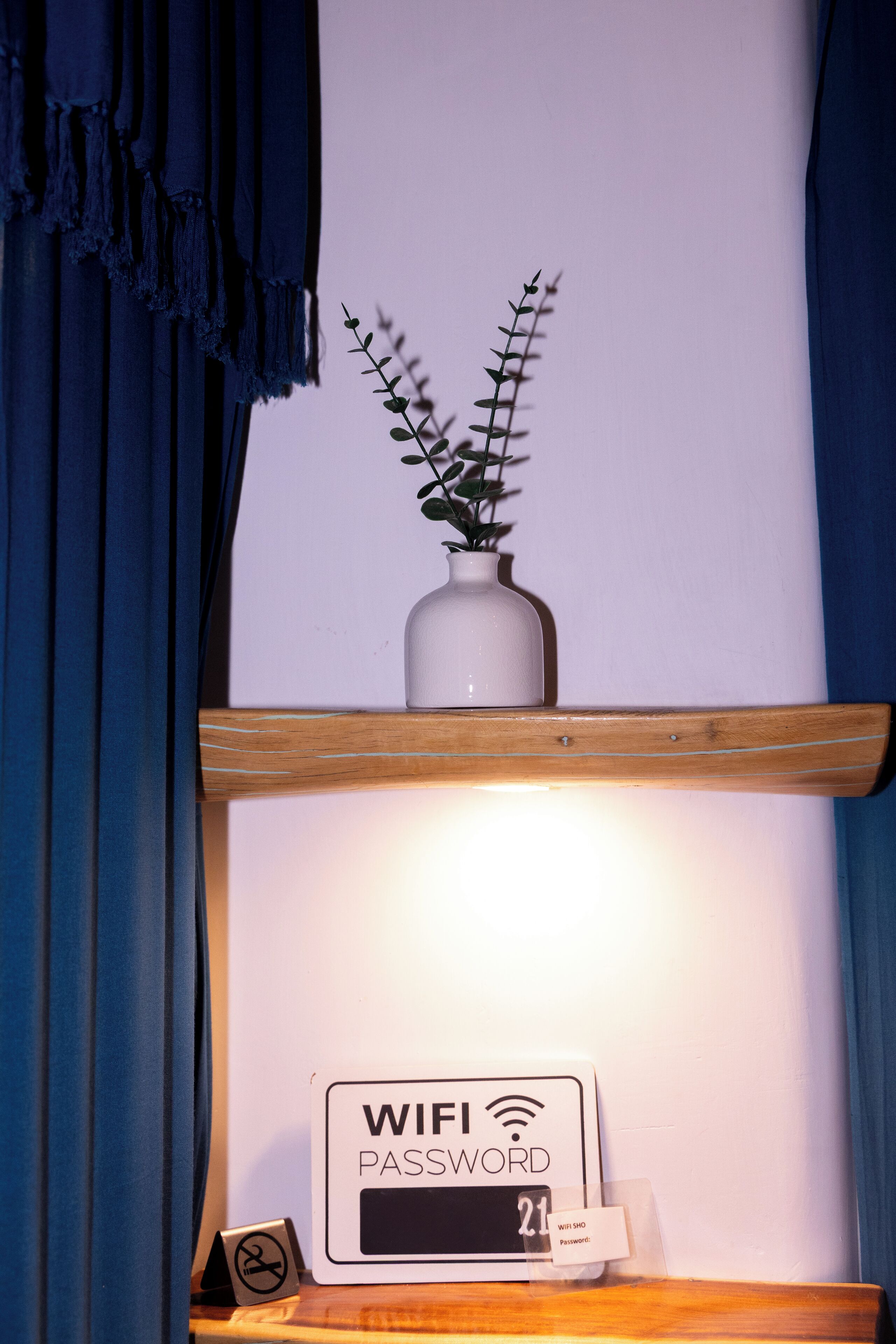 A room with a wooden shelf holding a white vase with a plant, a digital display with a WiFi password sign, and a small framed picture.