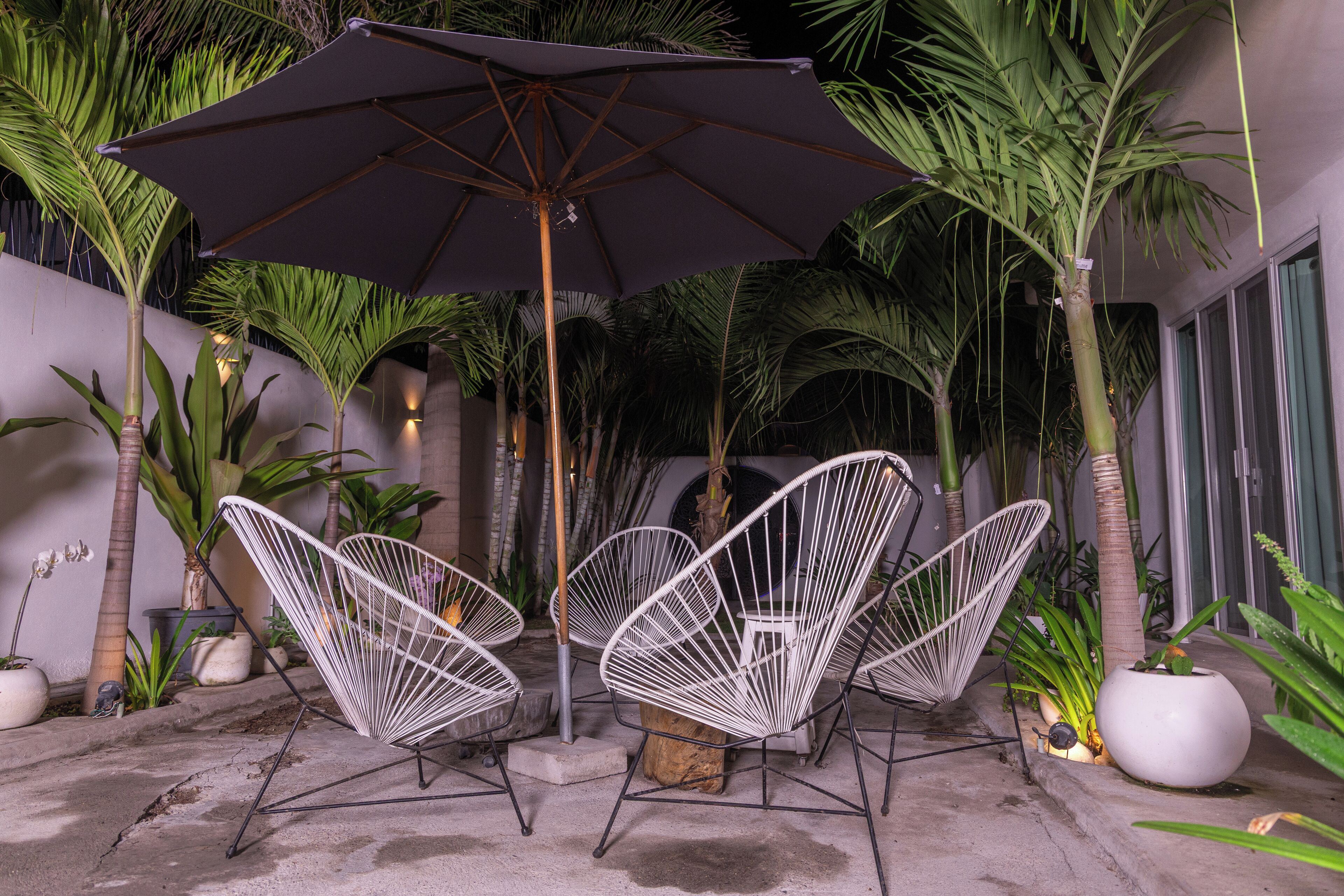 A patio area with two white wireframe chairs, a large dark umbrella, and various potted plants.
