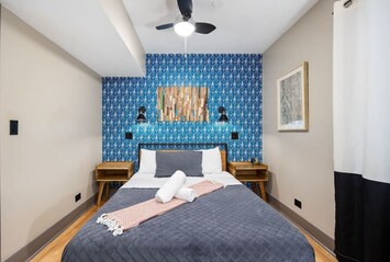 Cozy bedroom with a patterned wallpaper wall, a large mirror, a queen size bed with grey linens, a bedside table with a lamp and towels, and a window with white curtains.