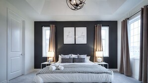 Ein modernes Schlafzimmer mit einem großen Bett, zwei Nachttischen mit Lampen und einer dunkel gestrichenen Wand mit zwei gerahmten Motivationssprüchen.
