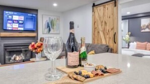 Een moderne woonkamer met een flatscreen televisie, een open haard en een eethoek met een glazen tafel, gedekt met snacks en een fles wijn.