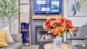 Een woonkamer met een flatscreen-tv aan de muur, waarop een streamingdienst te zien is, een open haard met een vaas bloemen, en een bank met kussens.