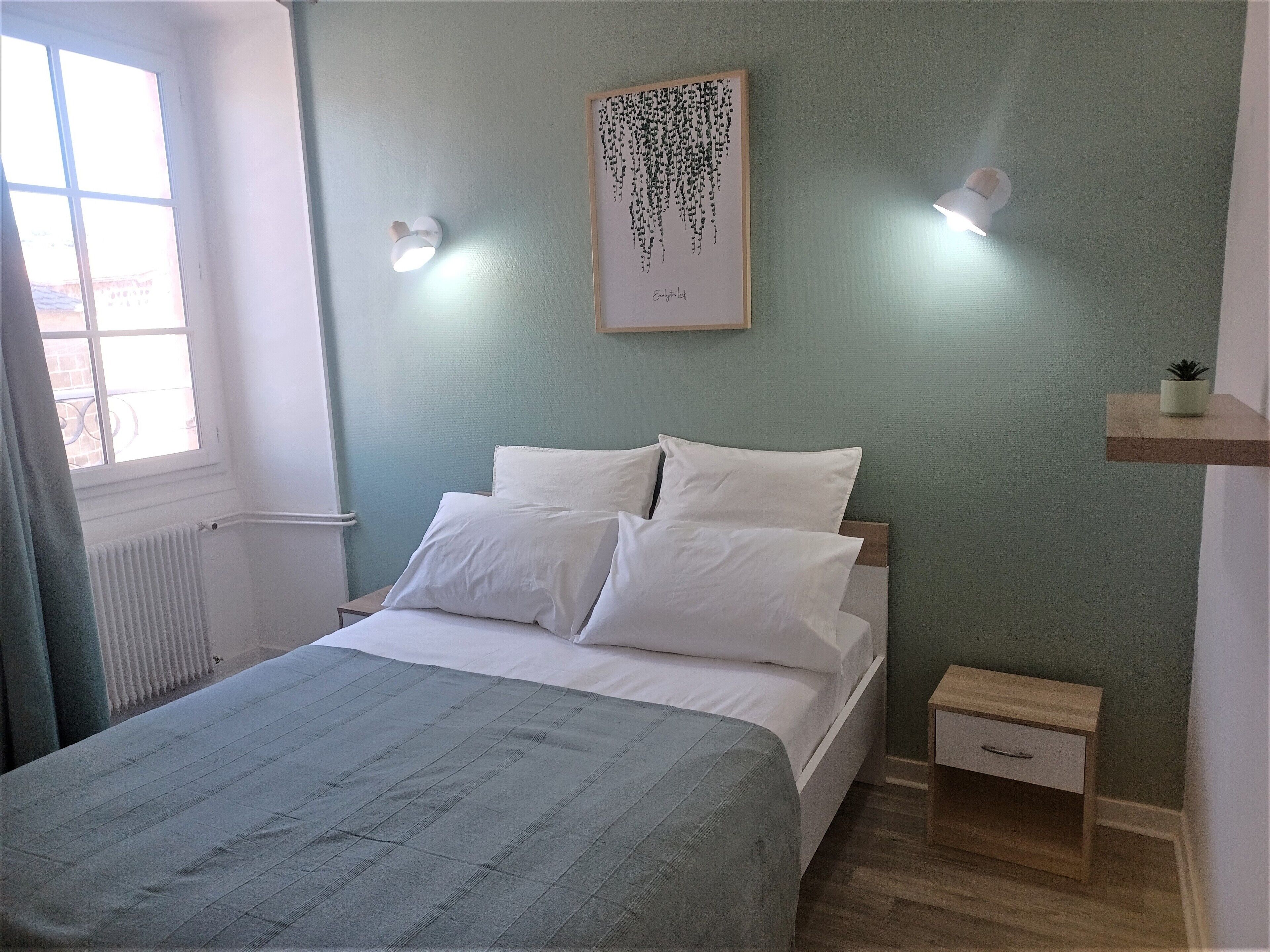 A neatly made bed with white linens, a small bedside table with a drawer, a wall-mounted light fixture, and a framed artwork above the bed.