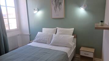 A neatly made bed with white linens, a small bedside table with a drawer, a wall-mounted light fixture, and a framed artwork above the bed.