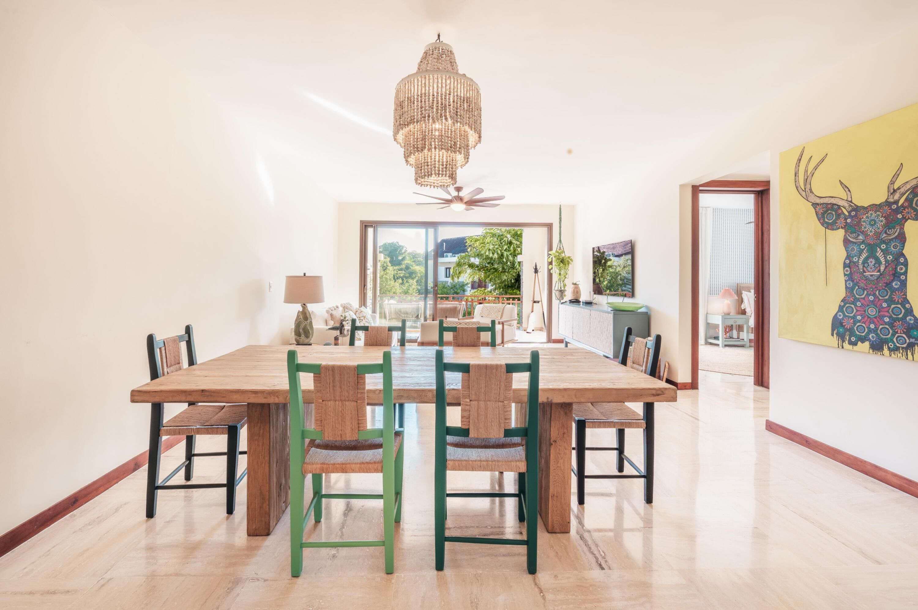 A spacious dining area with a large wooden table and green chairs, a chandelier, and a view of the outdoors.