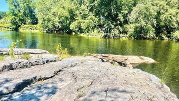Een natuurlandschap met een rivier, een rotsachtig voorplan en weelderige begroeiing op de achtergrond.