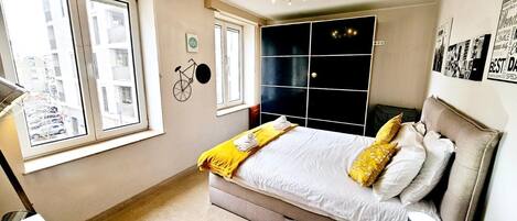 A bedroom with a bed, bedside table, lamp, and a view of the street through large windows.