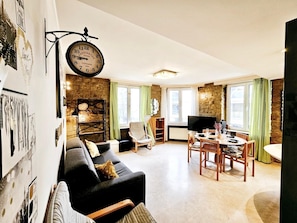 A cozy living room with a black sofa, a dining table with chairs, a television, and a large wall clock. - New 1 bedroom Flat in City Center  (Luxembourg City)