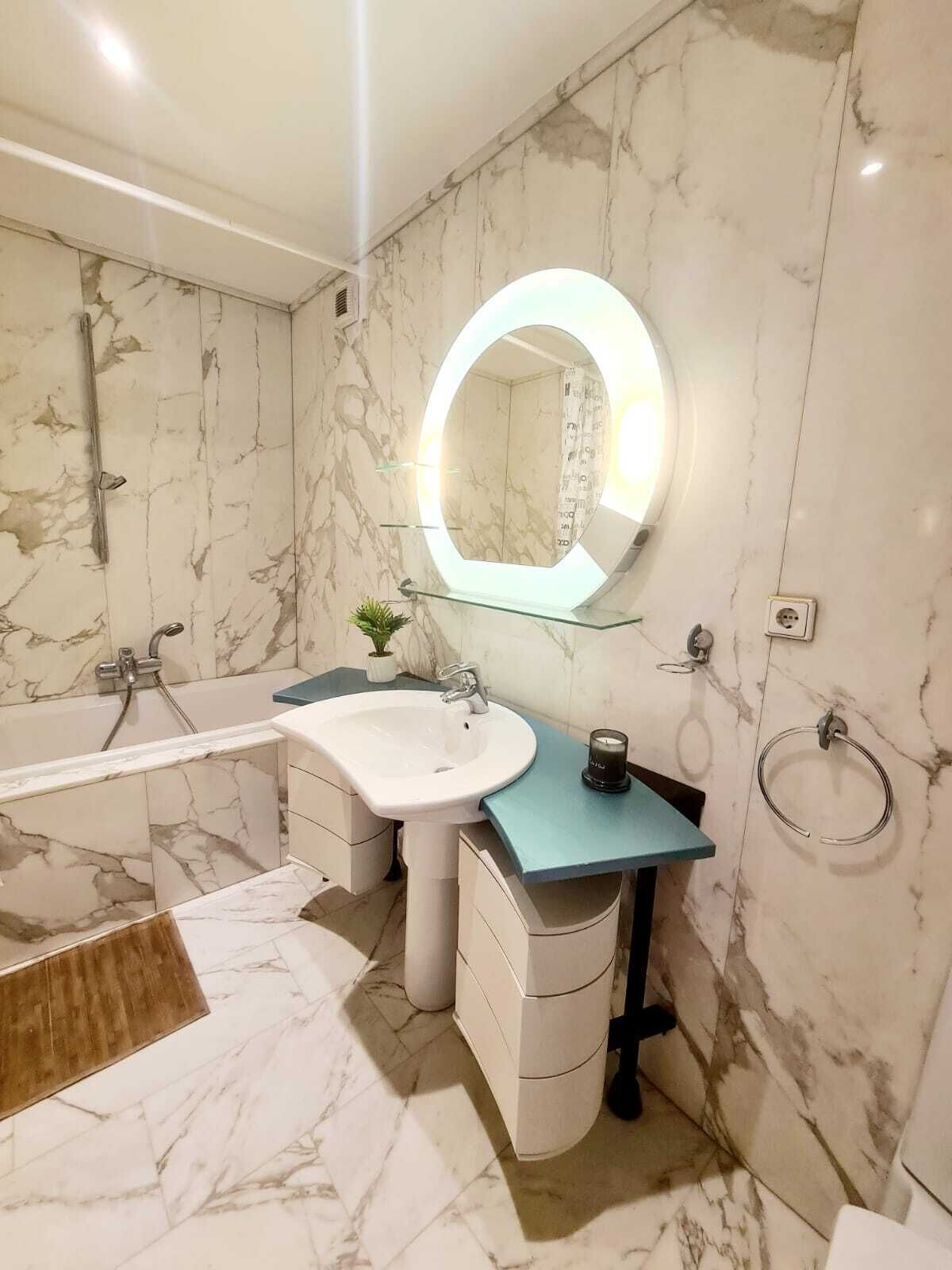 A modern bathroom with a marble floor, a large round mirror, a bathtub, a sink, and a toilet.