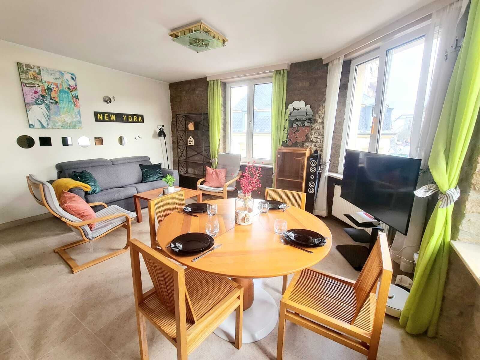 A modern living room with a dining area, a grey sofa, a wooden dining table set, and a TV on the right.