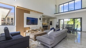 A spacious living area with a large grey sofa, a smaller black sofa, and a wooden coffee table. A flat-screen TV is mounted on the wall, and there is a dining area with a table and chairs visible in the background. - Brand new luxury villa located in a the most pristine gated community SOV
 (Sosúa)