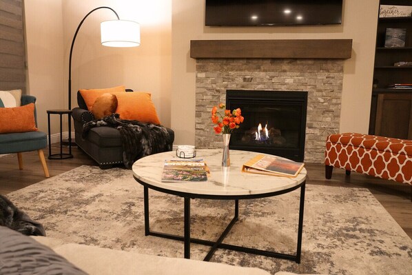 Una sala de estar con chimenea, una mesa de centro redonda con libros y un florero, un sofá con cojines naranja y una lámpara de pie.