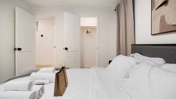 Ein Hotelzimmer mit einem ordentlich bezogenen Bett, einem Kleiderhaken mit einer Tasche und einer Tür zu einem Flur.