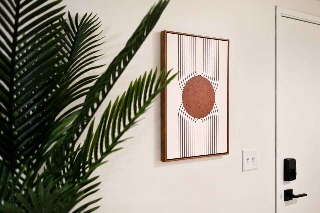 Ein gerahmtes Kunstwerk mit vertikalen Linien und einem zentralen Kreis an einer weißen Wand, neben einer Tür mit schwarzem Griff und einer Steckdose.