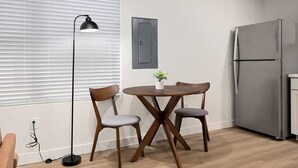 Ett litet kök med ett modernt kylskåp, ett matbord med två stolar, en golvlampa och ett fönster med persienner.