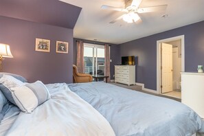 Un dormitorio con cama, cómoda, silla, televisor y puerta que da a un baño.