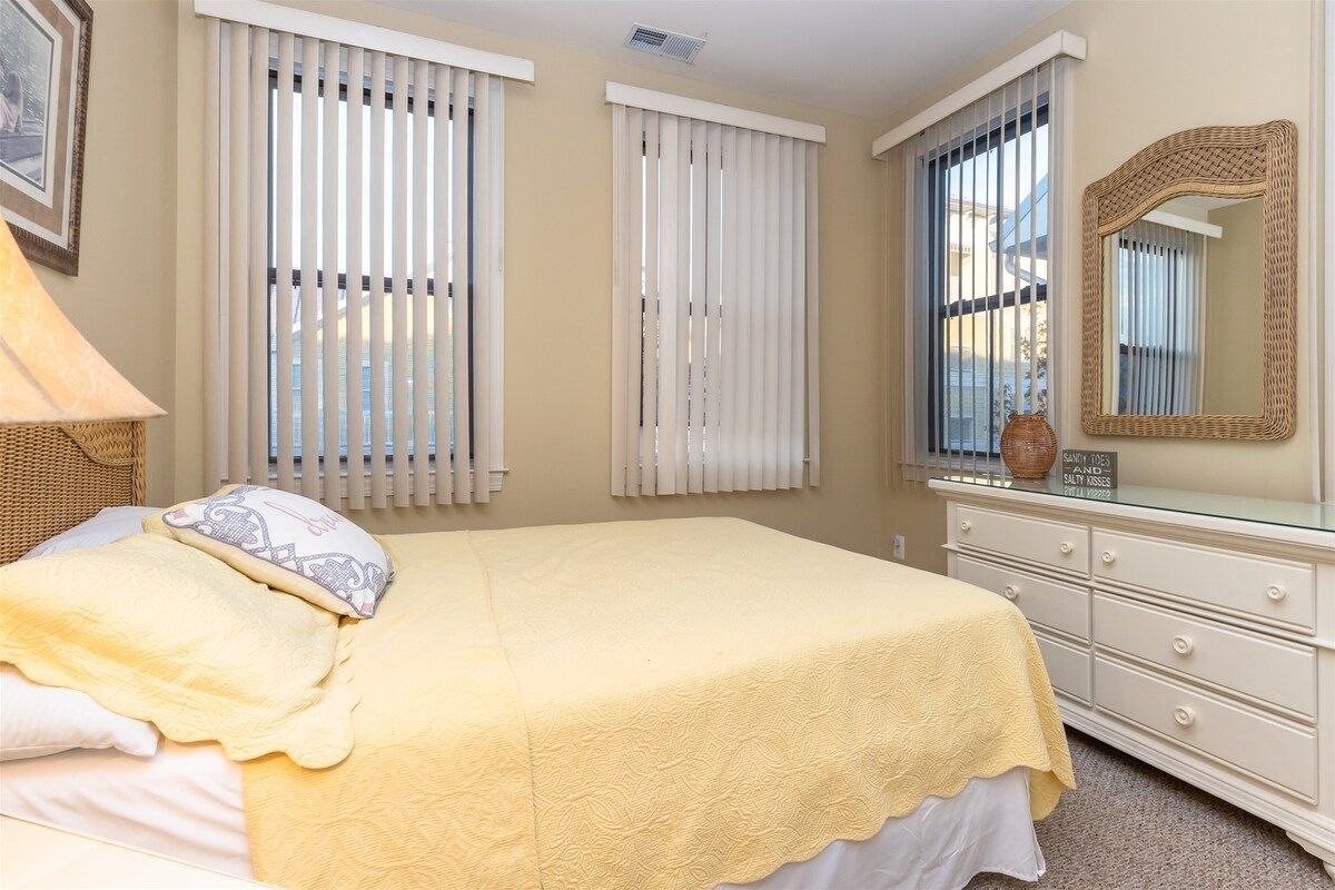 Une chambre à coucher comprenant un lit, une commode, un miroir et deux fenêtres équipées de stores.