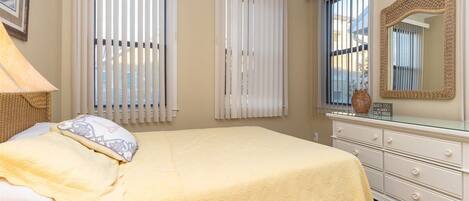 Une chambre à coucher comprenant un lit, une commode, un miroir et deux fenêtres équipées de stores.