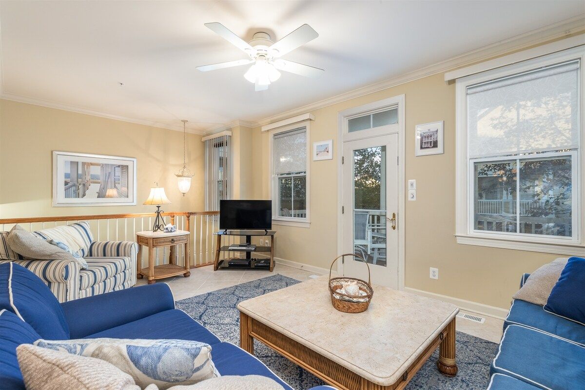 A living room with a television, a ceiling fan, a large window, and a door leading outside.