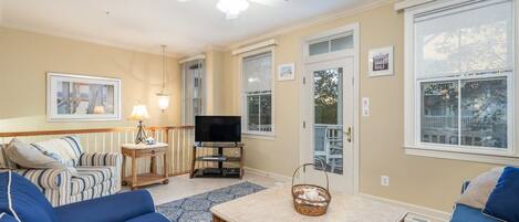 A living room with a television, a ceiling fan, a large window, and a door leading outside.