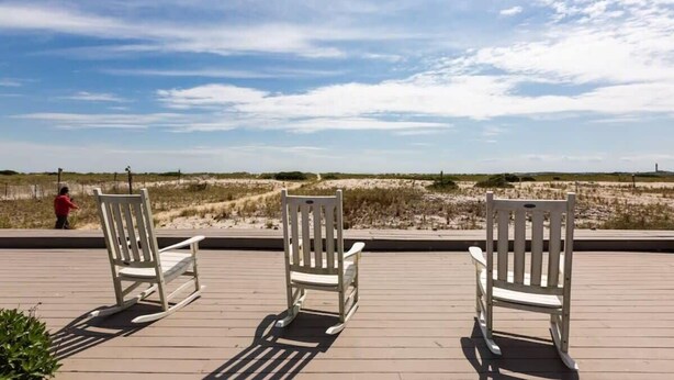 Een terras met drie schommelstoelen, gericht op een strand met duinen en een vuurtoren in de verte.