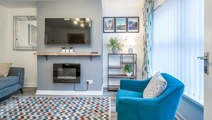 Una sala de estar con chimenea, televisor montado, un sillón azul, alfombra estampada y una ventana con persianas.