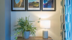 Una obra de arte con paisaje costero enmarco, una planta en maceta, una lámpara de mesa y un cortinaje estampado.