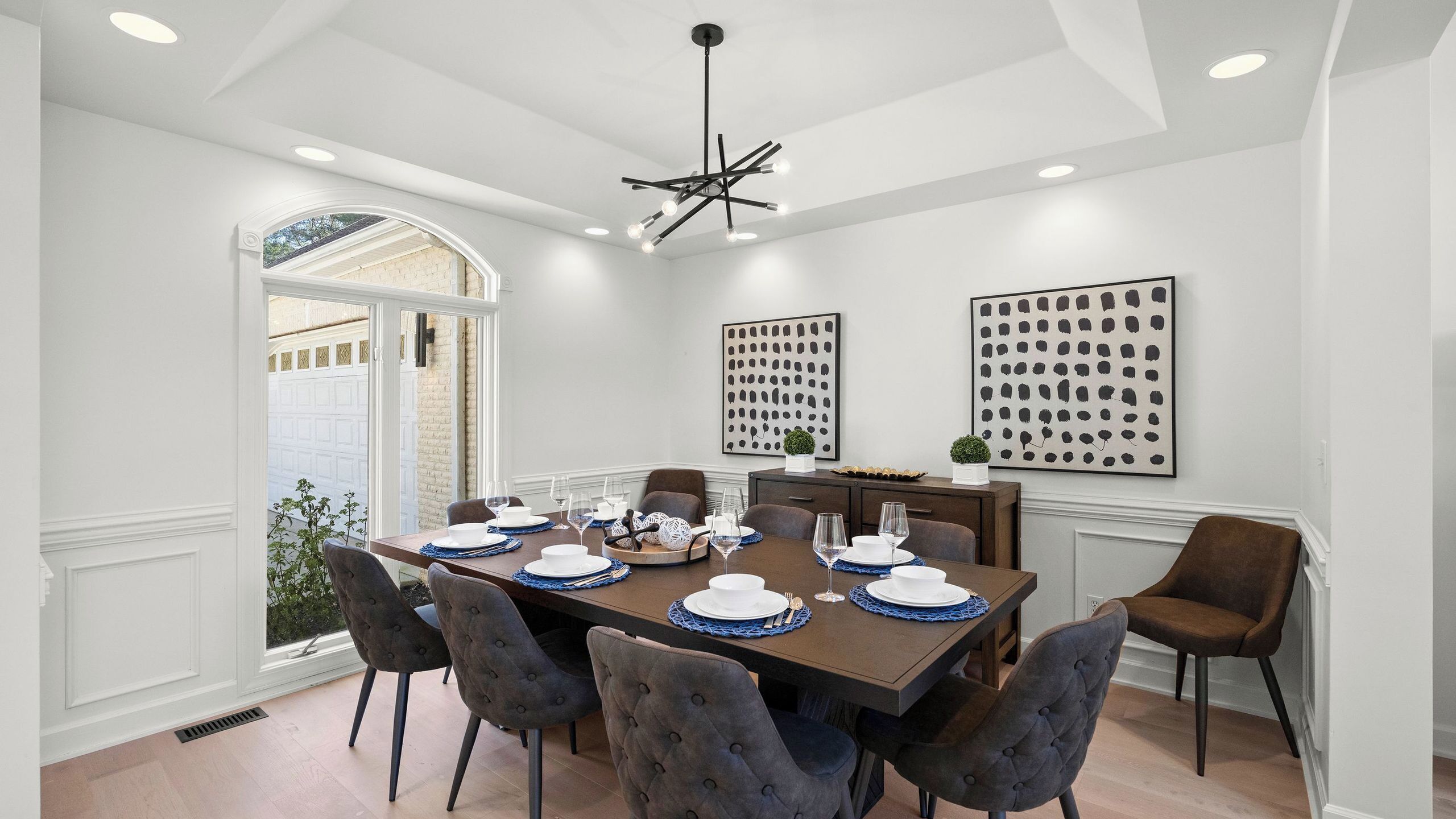 A dining room with a large table set for six, surrounded by six chairs, with two wall-mounted artworks and a ceiling light fixture.