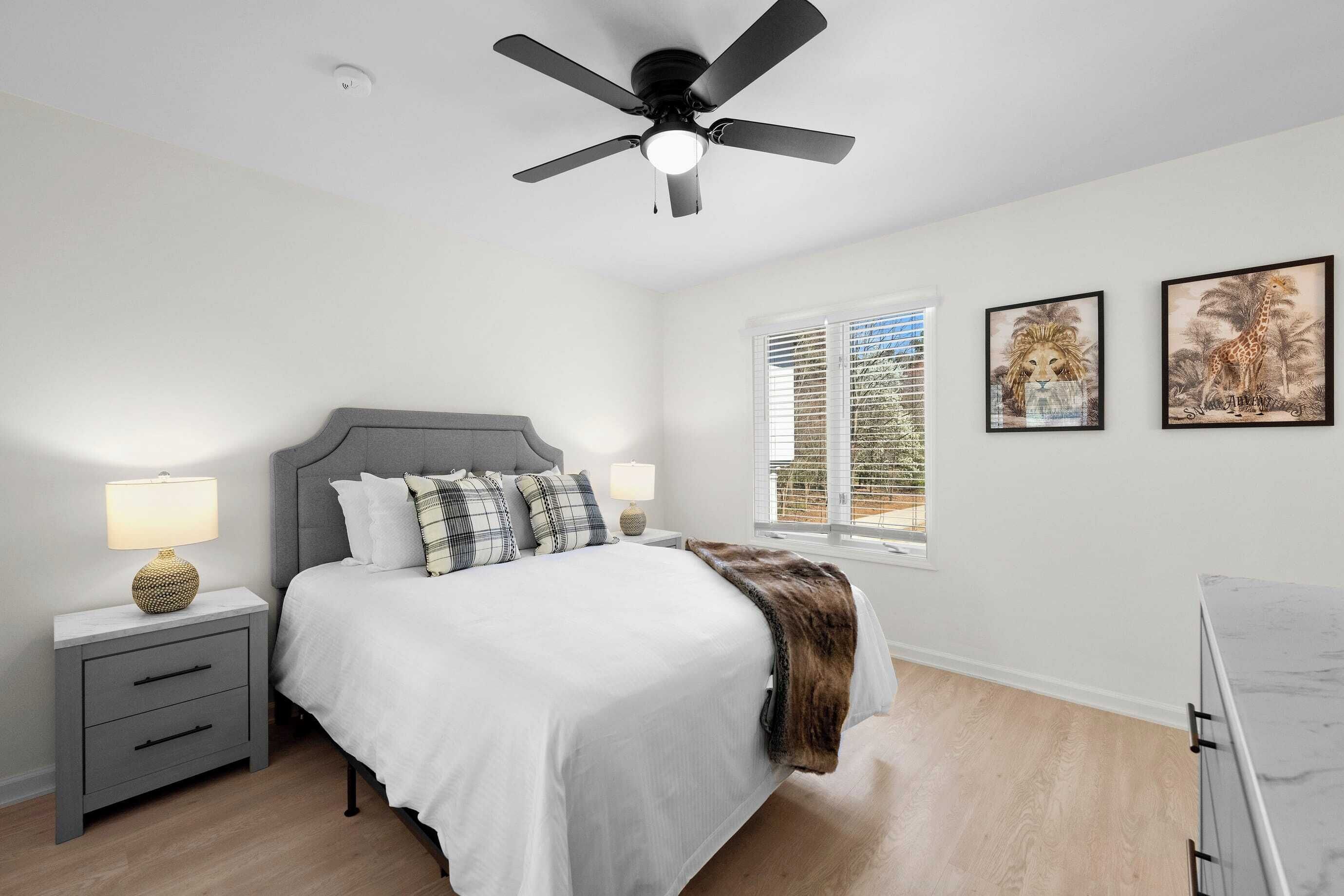 Une chambre à coucher comprenant un lit, une table de chevet, une lampe, un ventilateur de plafond, une fenêtre et deux tableaux encadrés accrochés au mur.